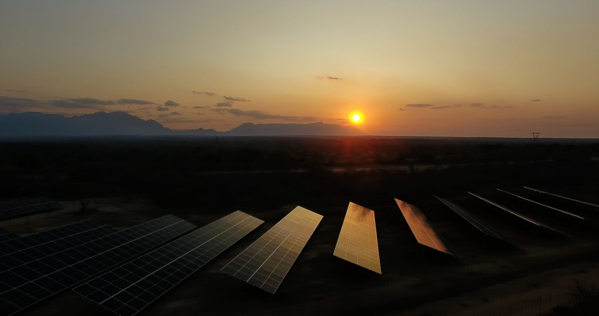 Aerial view of solar panel array at sunset, located alongside a remote highway with mountains in the background.