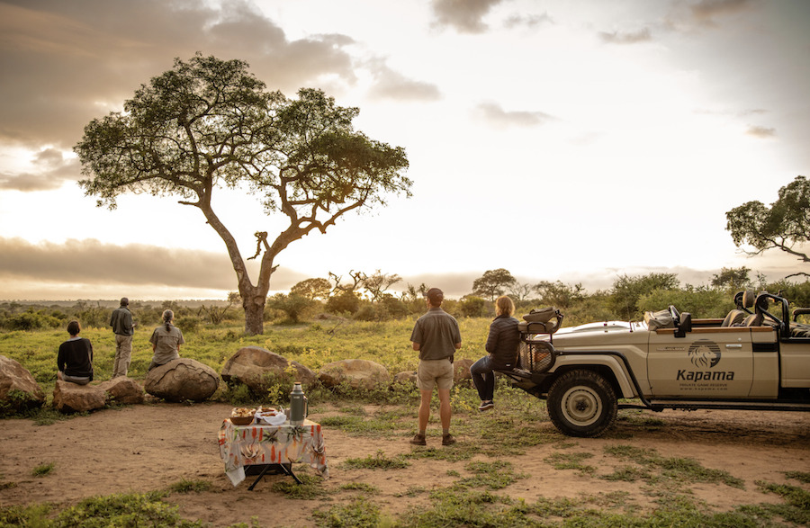 uests enjoying a serene sundowner break in the African bushveld with a Kapama Private Game Reserve safari vehicle parked nearby, overlooking the sunset and savanna landscape.